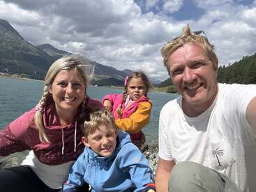 Eine glückliche vierköpfige Familie posiert an einem See mit Bergen im Hintergrund. Sie lächeln und genießen die Natur an einem sonnigen, teilweise bewölkten Tag.