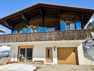 Das Bild zeigt ein charmantes Chalet mit einem Holzbalkon, blauen Fensterläden und einem Satteldach. Auf dem Boden liegt Schnee, und der Himmel ist strahlend blau.
