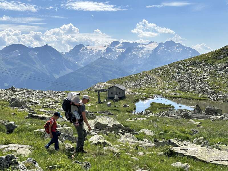 Ein Mann und zwei Kinder wandern durch eine felsige, grasbewachsene Berglandschaft. Unter strahlend blauem Himmel sind schneebedeckte Gipfel, ein kleiner Teich und eine Hütte zu sehen.