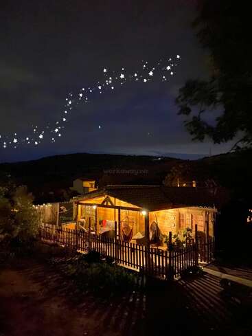 L'image représente une maison en bois confortable avec un porche et une clôture, illuminée la nuit, sur fond de ciel étoilé et entourée d'arbres et de collines.