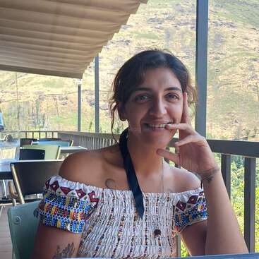 Una mujer sentada en un café al aire libre sonríe con la mano apoyada en la cara. Lleva un top estampado sin hombros. Las montañas forman el fondo escénico.