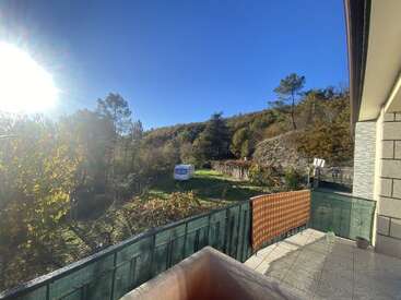 A bright, sunny morning view from a tiled balcony, overlooking a green yard with a caravan, surrounded by lush trees and distant forested hills. Peaceful atmosphere.