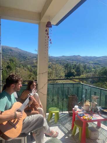 A man plays guitar while a woman listens on a sunny balcony overlooking beautiful green mountains. A small table with items and stools completes the scene.