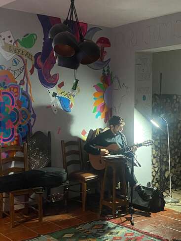 A person sits on a stool playing guitar, singing into a microphone. Artistic, colorful murals and warm lighting create a cozy, creative indoor atmosphere with chairs and decorations.