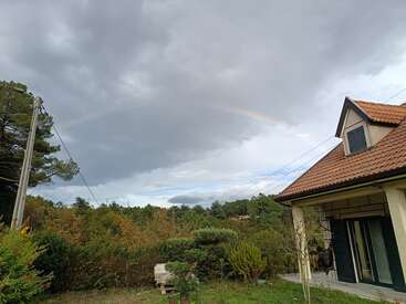 A cozy house with a red-tiled roof sits near lush green bushes and trees. Overhead, a faint rainbow arcs across the cloudy, dramatic sky.
