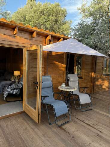 A imagem mostra uma cabana de madeira aconchegante com uma porta aberta, convidando a uma atmosfera serena, com duas cadeiras, uma mesa e um guarda-chuva branco no deck externo.
