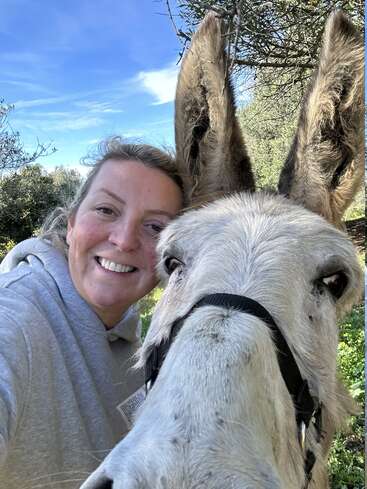A smiling woman in a grey hoodie takes a close-up selfie with a friendly donkey outdoors. They both look happy under a bright blue sky.