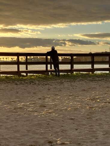 A person leans on a wooden fence, gazing at the water during sunset. The sky is dramatic, with clouds and golden sunlight illuminating the sandy foreground.