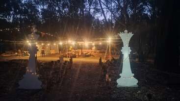 Un caprichoso evento al aire libre, con decoraciones de piezas de ajedrez gigantes, luces brillantes, asientos acogedores y un ambiente cálido y acogedor rodeado de altos árboles al atardecer.