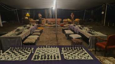 La imagen muestra una carpa con varios tableros de ajedrez colocados sobre largas mesas, cojines para sentarse, cómodos sofás, una iluminación tenue y un ambiente relajado.