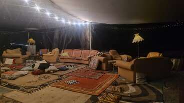 La imagen muestra una acogedora zona de estar al aire libre con sofás, sillones, alfombras y muchos cojines. Las luces de cuerda y una lámpara de pie crean un ambiente cálido y acogedor.