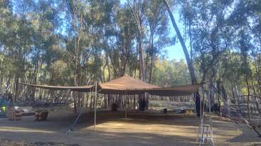 Una gran tienda de campaña marrón está instalada en un claro del bosque, rodeada de árboles altos. Debajo y alrededor se ven sillas de acampada, material y una escalera.