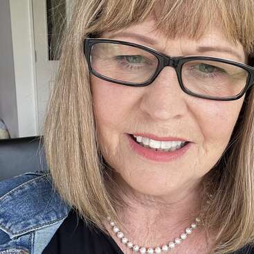 A woman with straight blonde hair, glasses, and a pearl necklace is smiling. She is wearing a denim jacket and black top, photographed in natural light indoors.