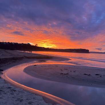 A stunning sunset illuminates the sky with vibrant orange and purple hues, reflected on calm water. Silhouetted figures walk along a peaceful sandy beach.