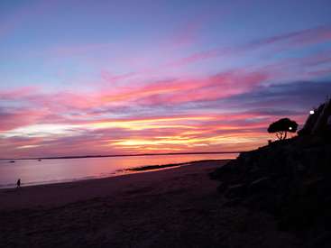 La imagen representa una serena escena playera al atardecer, con un cielo vibrante, aguas tranquilas y una figura solitaria paseando por la orilla, en un ambiente tranquilo.