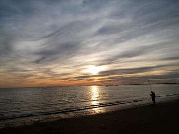 La imagen representa una serena escena playera al atardecer, con una persona pescando en la orilla y una barca visible en el agua.