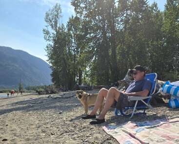 Um homem está sentado numa cadeira de praia ao lado do seu cachorro, relaxando numa praia ensolarada de seixos, com árvores, montanhas e pessoas ao fundo, curtindo a natureza.