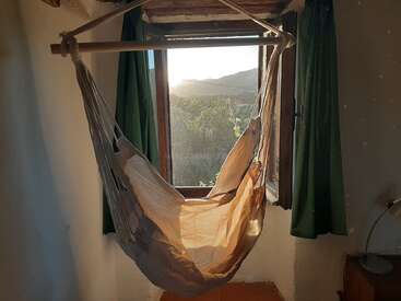 La imagen representa una serena escena de una silla hamaca suspendida frente a una ventana, con una vista de los árboles y las colinas del exterior, bañados por la cálida luz del sol.