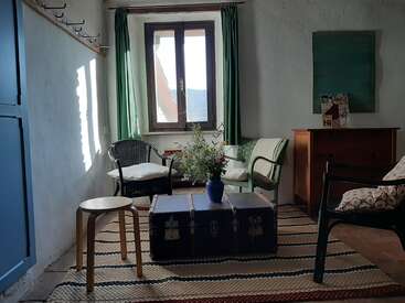 La imagen representa una acogedora sala de estar con una ventana, en la que hay un sofá, dos sillas, una mesa de centro y una alfombra, que desprenden un ambiente cálido y acogedor.