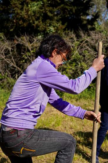 La imagen muestra a una persona vestida con camisa morada y vaqueros, agarrando un poste de madera con ambas manos, sobre un fondo de árboles y hierba.