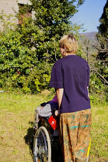 La imagen muestra a una mujer de pelo corto y rubio de pie en una zona de césped, de espaldas a la cámara, con una silla de ruedas a su lado.