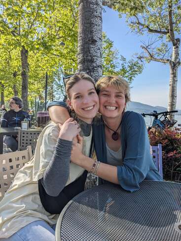 Dos mujeres sentadas en una mesa, abrazadas, con árboles, un lago y una montaña de fondo, rodeadas de otras personas y bicicletas.