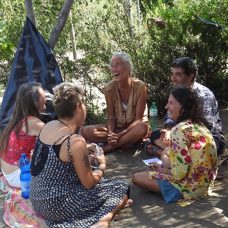 La imagen muestra a un grupo de cinco personas sentadas en círculo en el suelo, rodeadas de árboles y arbustos, enzarzadas en una conversación.