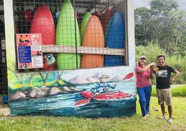 Two people stand smiling in front of a mural showing a kayaker. Behind them are colorful kayaks stored vertically. Posters about mental health are visible.