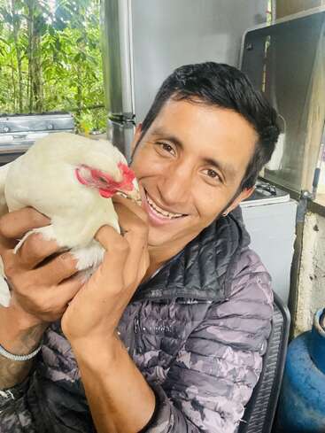A smiling man in a gray jacket holds a white chicken affectionately indoors, with greenery visible outside the window behind him, creating a warm, joyful moment.