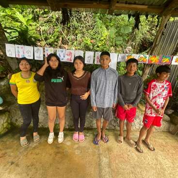 Six children stand in a row outdoors under a shelter, with colorful drawings and paper art displayed on a string behind them. Lush greenery surrounds them.