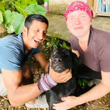 Two people, one smiling brightly and the other wearing a red bandana, pose happily outdoors with a black dog between them, surrounded by greenery and books.