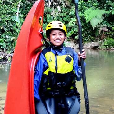 A smiling child stands in shallow water, holding a paddle and red kayak. Wearing a yellow helmet and life jacket, surrounded by lush green forest foliage.