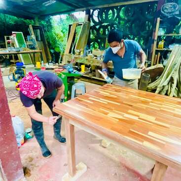Two people in a workshop are carefully varnishing a wooden table. Various tools, wood pieces, and materials can be seen around, suggesting active woodworking and craftsmanship.