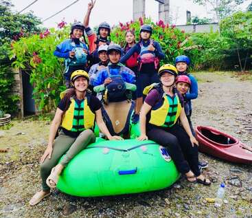 A group of people wearing safety helmets and life jackets pose together, smiling, around a green inflatable raft and kayaks, surrounded by greenery and bright flowers.