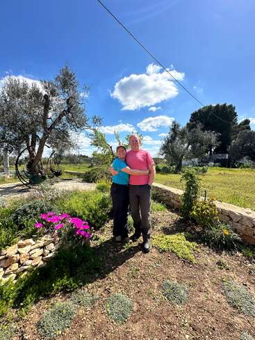 A couple stands arm in arm in a sunny garden, surrounded by greenery, blooming pink flowers, and olive trees, under a bright blue sky.