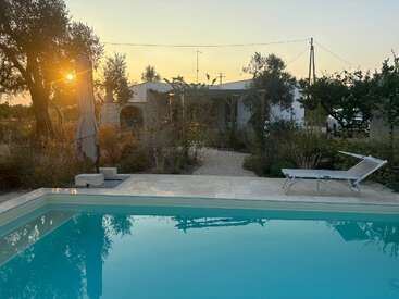 A peaceful backyard scene at sunset, featuring a clear swimming pool, a lounge chair, an umbrella, lush greenery, and a quaint white house in the background.