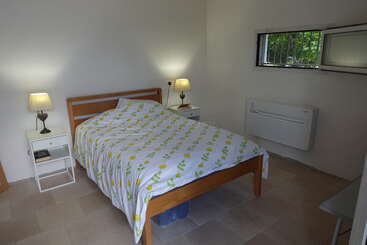 This is a simple, tidy bedroom with a wooden bed, patterned bedding, two bedside tables with lamps, a small window, and a wall-mounted air conditioner.