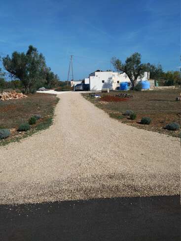 Une allée de gravier mène à une maison rurale blanche entourée d'arbres et de terres ouvertes. Des réservoirs d'eau bleue se trouvent à proximité, sous un ciel bleu clair.