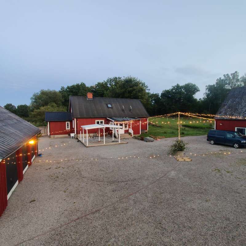 La imagen representa una serena escena rural con un granero rojo de tejado oscuro, rodeado de un camino de grava y adornado con cuerdas de luces, sobre un cielo nublado.