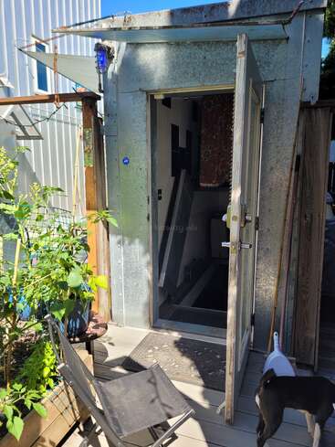 Ein kleiner Metallschuppen mit einer offenen Tür gibt den Blick auf einen Innenraum mit Treppe frei. Draußen gibt es einen Stuhl, ein paar Topfpflanzen und einen Hund, der herumschnüffelt.