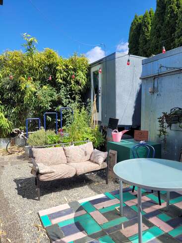 Auf einer gemütlichen Terrasse im Hinterhof stehen ein gemustertes Sofa, ein Glastisch, bunte Fliesen, üppige Grünpflanzen, Lichterketten, eine rosa Gießkanne und ein sonniger blauer Himmel.