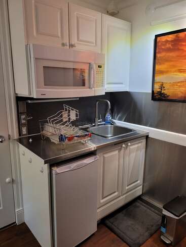 Dieses Bild zeigt eine kompakte Küchenzeile mit weißen Schränken, einer Mikrowelle, einem kleinen Kühlschrank, einer Arbeitsplatte und Spüle aus Edelstahl, einem Geschirrkorb, einem Sonnenuntergangskunstwerk und einem Mülleimer.