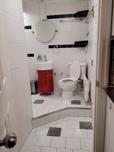 Dies ist ein modernes Badezimmer mit weißen Fliesen, schwarzen Akzenten, erhöhtem Boden, Toilette, rundem Spiegel, rotem Waschtisch, Regalen, Handtuchhaltern und einem kleinen Mülleimer.