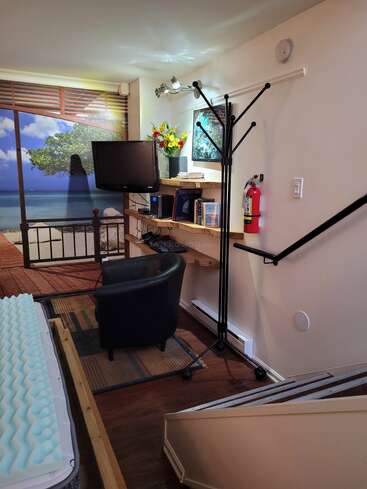 Ein gemütliches Zimmer mit einem schwarzen Stuhl, einem Fernseher, einem Bücherregal, Blumen, einem Kleiderständer und einem Wandbild am Strand. Eine Matratze mit Schaumstoffauflage, ein Feuerlöscher und eine Treppe sind auch dabei.