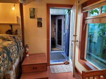 Ein gemütliches Zimmer mit geblümter Bettwäsche, offener Tür, Fenster mit Jalousien, Feuerlöscher, Holzstufen, Wandkunst und sichtbarem Grün draußen, das eine einladende Atmosphäre schafft.