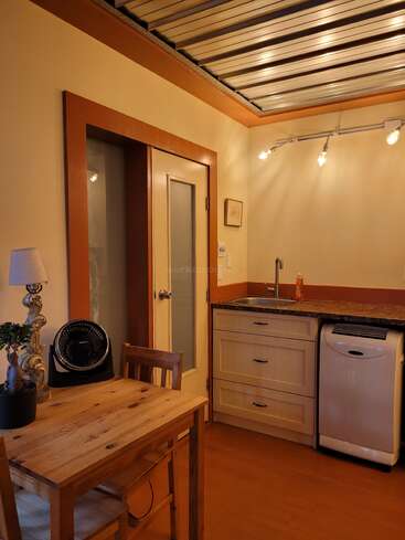 Diese gemütliche Küchenzeile ist mit Holzmöbeln, einem kleinen Esstisch, einer Lampe, einer Topfpflanze, einem Ventilator, einem kompakten Waschbecken und einer Deckenbeleuchtung ausgestattet.