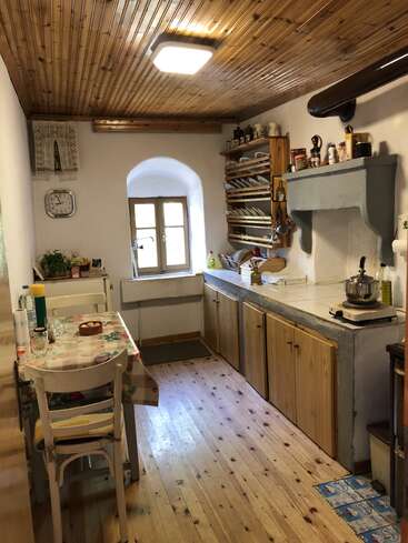 This image shows a cozy, rustic kitchen with wooden cabinets, a small table, dishes on shelves, a window, wooden ceiling, and warm, inviting atmosphere.