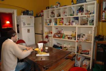 The image depicts a man sitting at a table in a kitchen, surrounded by a refrigerator, shelves, and various food items, with a lamp in the background.