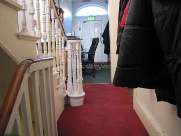 The image depicts a hallway with a red carpet, white banister, and coat rack, leading to a doorway with a white door and arched window.