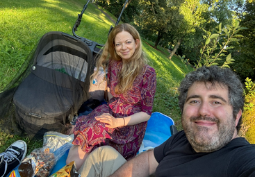 A couple enjoys a sunny picnic in a lush green park, sitting on a blanket next to a stroller, snacks, and wearing comfortable, casual clothing.